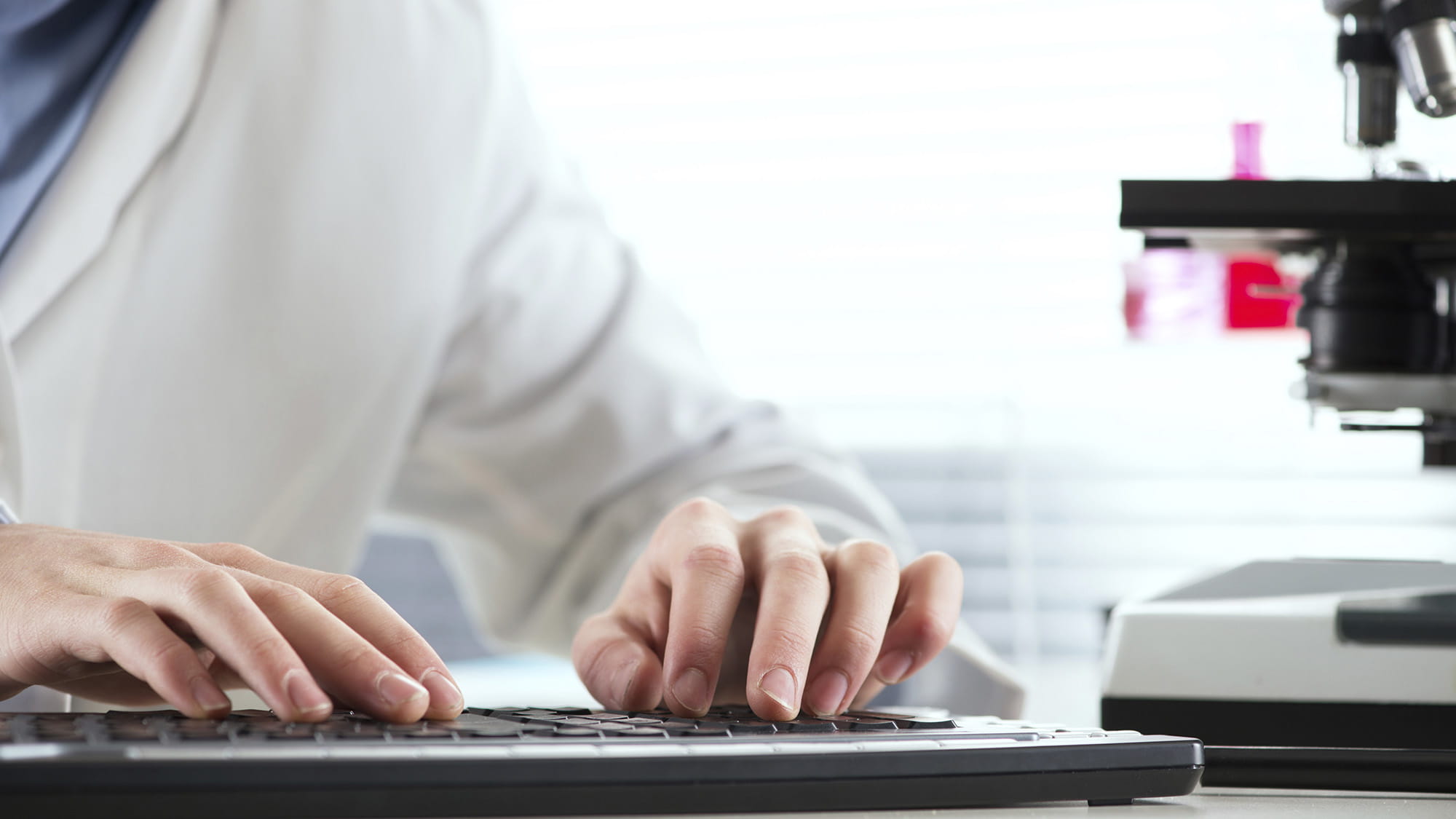 Researcher in white coat at desk with hands on keypad, microscope cut off to the right