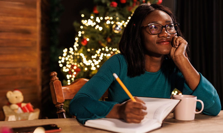 Happy young woman sitting at home office desk, contemplating her New Year's resolutions and writing them in a diary.